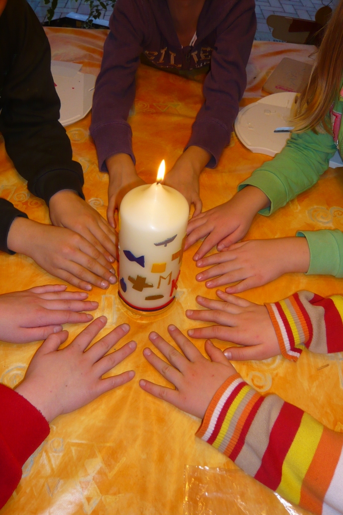 Kinderhände bilden liegend auf dem Tisch einen Kreis, in der Mitte steht die Kommuniongruppenkerze