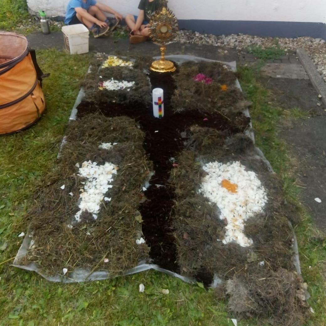 die Kinder haben einen kleinen Fronleichnamsteppich gestaltet, in der Mitte ein schwarzes Kreuz aus Blumenerde, darauf eine Kerze, aus weißen Blüttenblättern wurde ein Fisch gelegt.