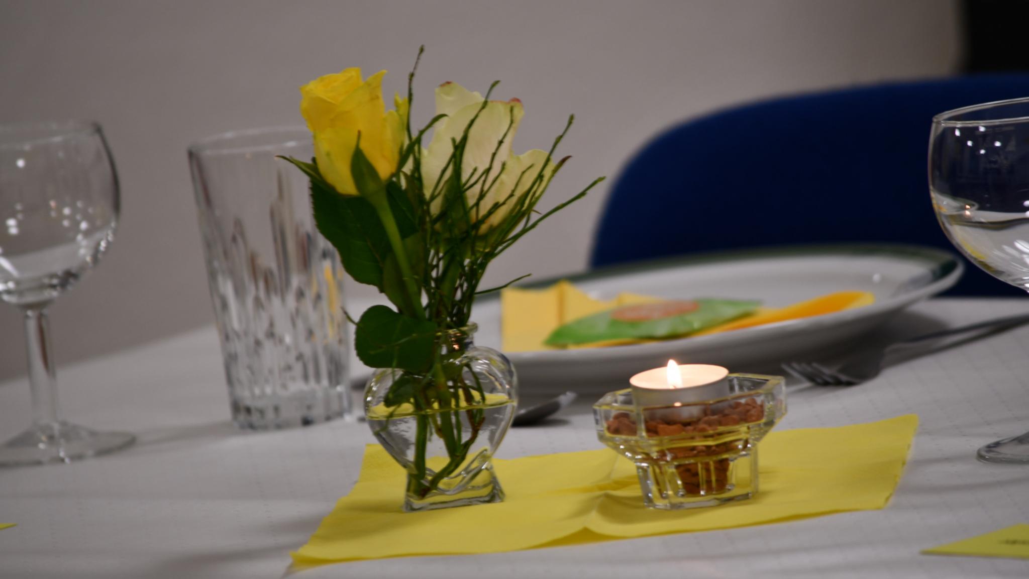Gelbe Rose in Vase, Teelicht und gedeckter Teller auf weißem Tisch mit gelber Serviette.